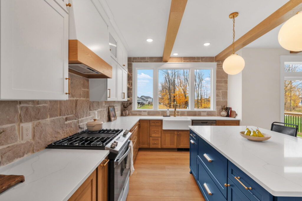 Spacious kitchen with a blue island, wood cabinetry, stone backsplash, exposed ceiling beams, and large windows overlooking autumn trees.