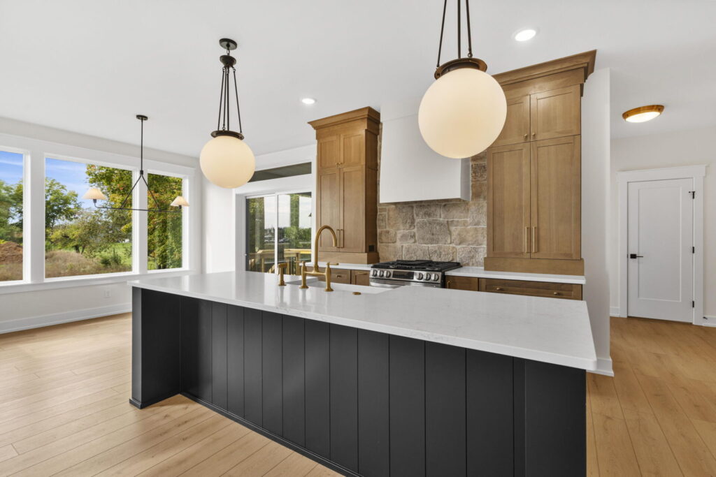 Modern kitchen with a long black island, round globe pendant lights, wood cabinetry, and stone backsplash with large windows bringing in natural light