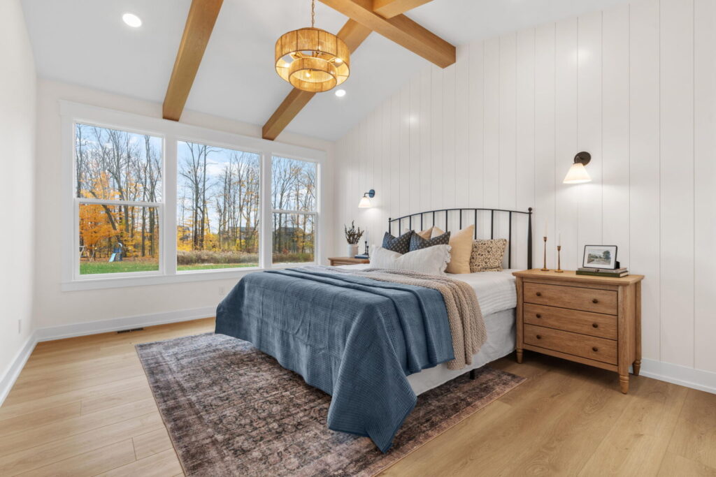Peaceful bedroom with a vaulted ceiling and wood beams, large windows overlooking fall trees, a metal bed frame, and wood nightstands.
