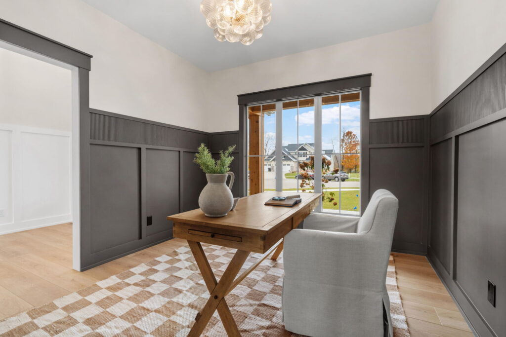 Cozy home office with dark gray wainscoting, light walls, a wood desk, plush armchair, and large windows facing a neighborhood street.