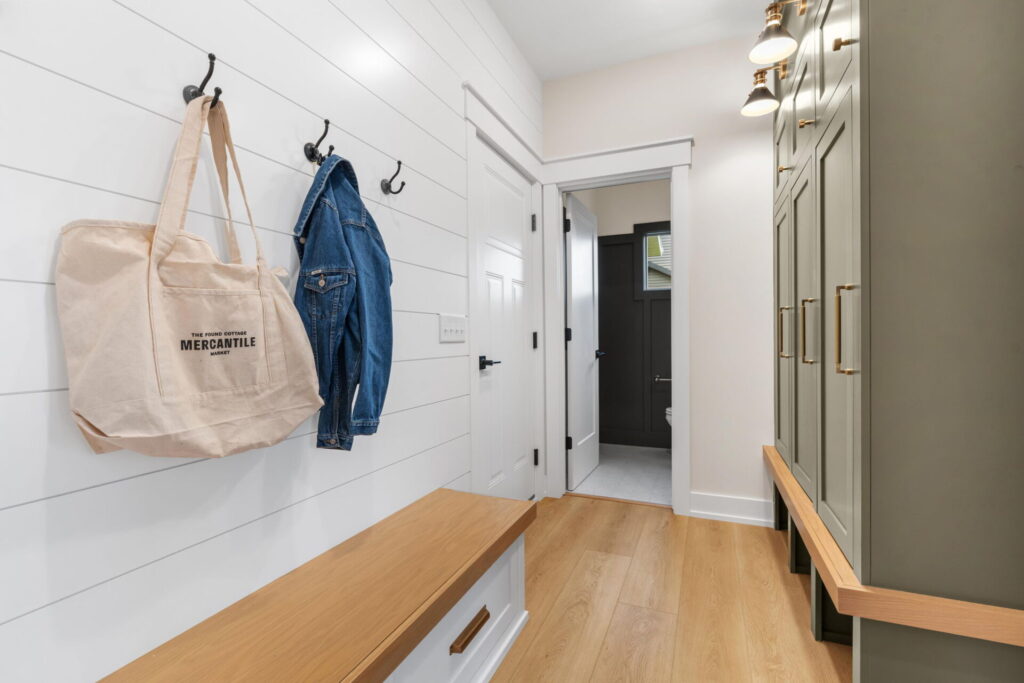 Bright mudroom with shiplap walls, wood bench seating, black hooks holding a tote bag and denim jacket, and tall sage-green built-ins with brass hardware.