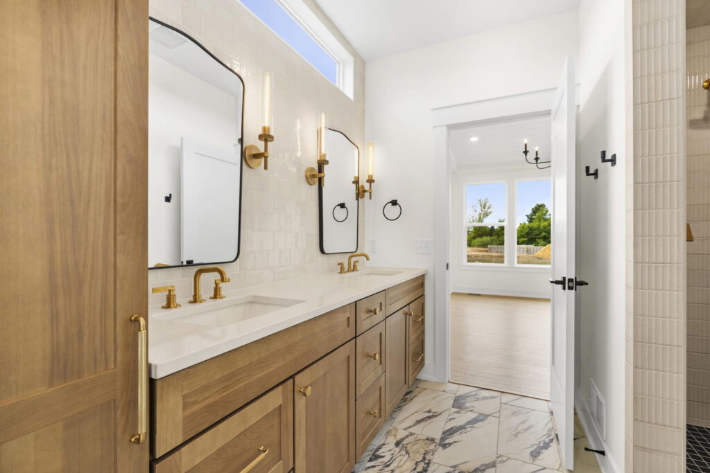 Warm, modern bathroom featuring a double vanity with wood cabinets, brass fixtures, sculptural sconces, and a doorway opening to a sunlit bedroom.